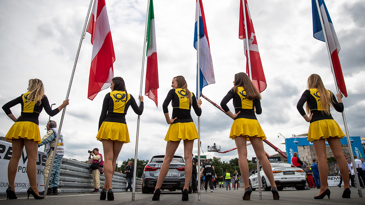 Five grid girls in short black and yellow dresses stand in a line holding flags from different countries at the start of a car race