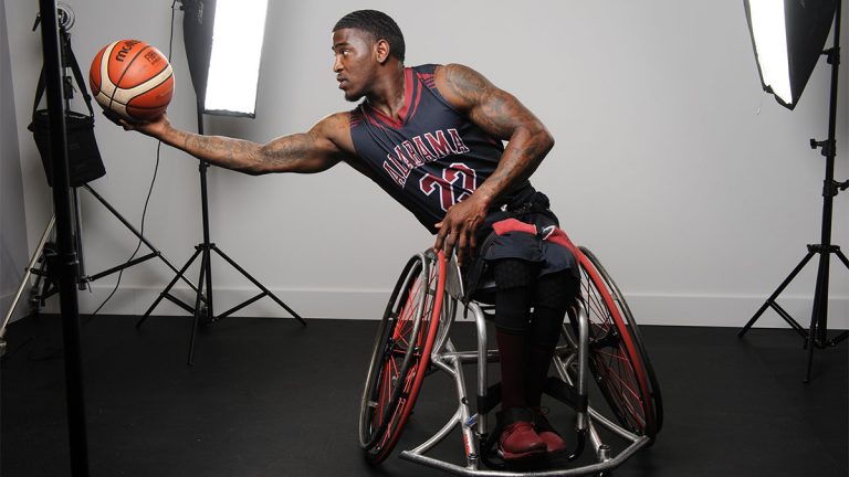Dequel Robinson in University of Alabama basketball jersey sitting in wheelchair with outstretched arm holding basketball